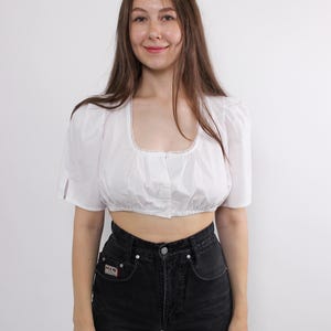 May include: White crop top with a square neckline and short, puffed sleeves. The top has a button-up front and is paired with high-waisted black jeans. The neckline is trimmed with a delicate lace detail. The outfit is simple and classic.
