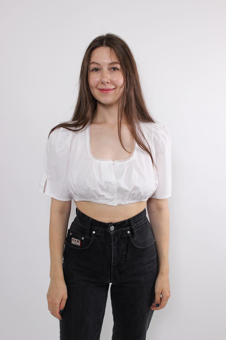 May include: White crop top with a square neckline and short, puffed sleeves. The top has a button-up front and is paired with high-waisted black jeans. The neckline is trimmed with a delicate lace detail. The outfit is simple and classic.