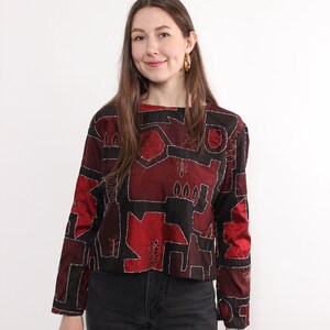 May include: A long-sleeved, cropped top with a unique geometric design in shades of red and black. The top features silver stitching outlining the shapes and is paired with black jeans. The top has a round neckline and button details.