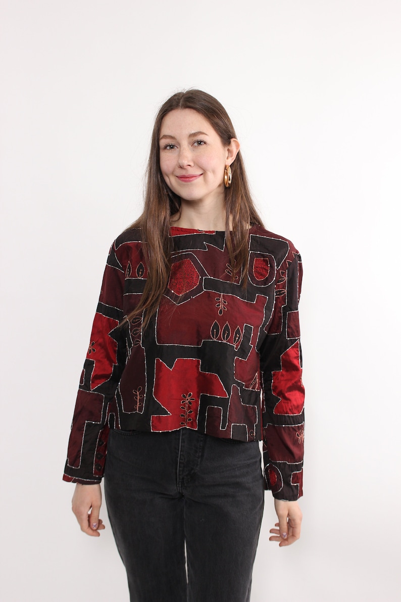 May include: A long-sleeved, cropped top with a unique geometric design in shades of red and black. The top features silver stitching outlining the shapes and is paired with black jeans. The top has a round neckline and button details.