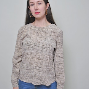 Puede incluir: Una mujer con una blusa de manga larga con estampado marrón y blanco, cuello redondo y pantalones vaqueros azules.