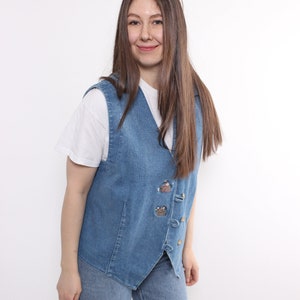 Op de afbeelding: Een vrouw draagt een blauwe denimvest met geborduurde details en een wit T-shirt. Ze draagt ook een blauwe jeans.