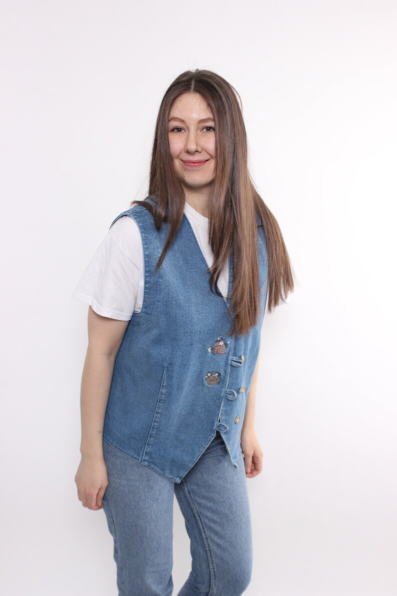 Op de afbeelding: Een vrouw draagt een blauwe denimvest met geborduurde details en een wit T-shirt. Ze draagt ook een blauwe jeans.