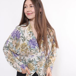 May include: A floral print blouse with a V-neckline. The blouse features a pattern of purple, green, and beige flowers on a cream background. The long-sleeved top is made of a lightweight fabric.