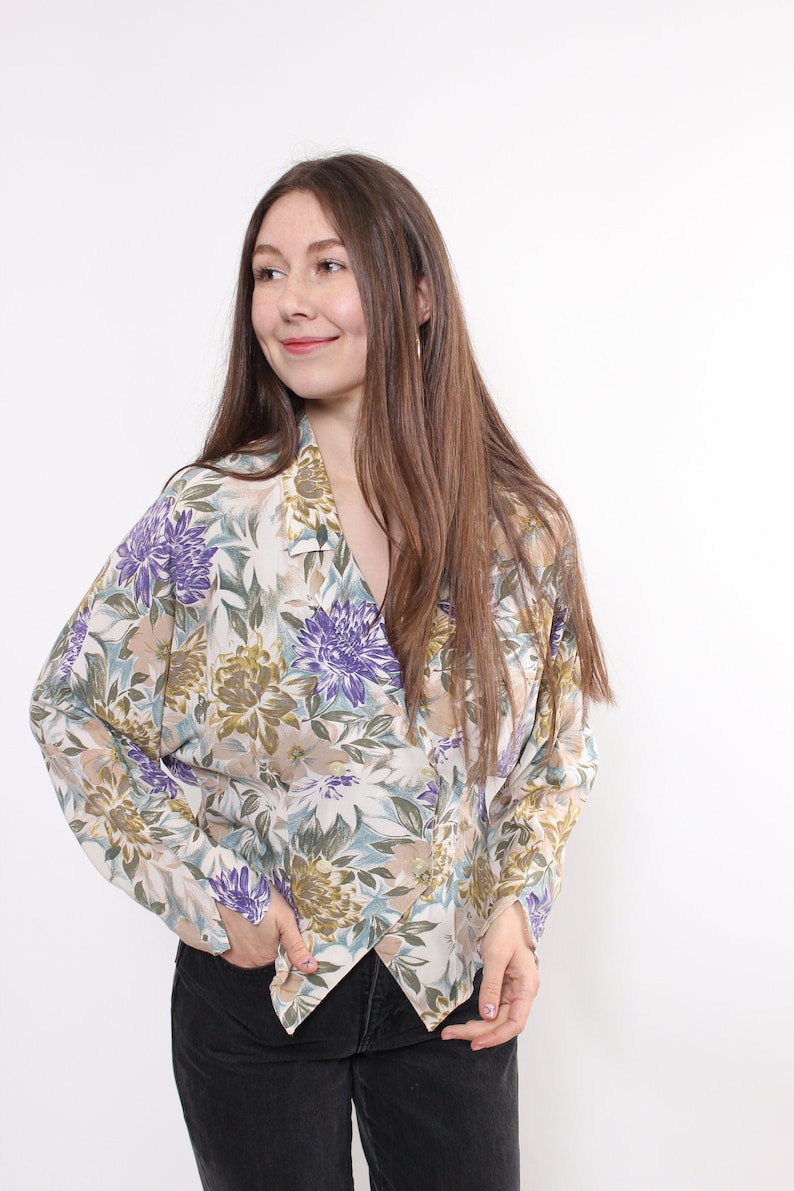 May include: A floral print blouse with a V-neckline. The blouse features a pattern of purple, green, and beige flowers on a cream background. The long-sleeved top is made of a lightweight fabric.