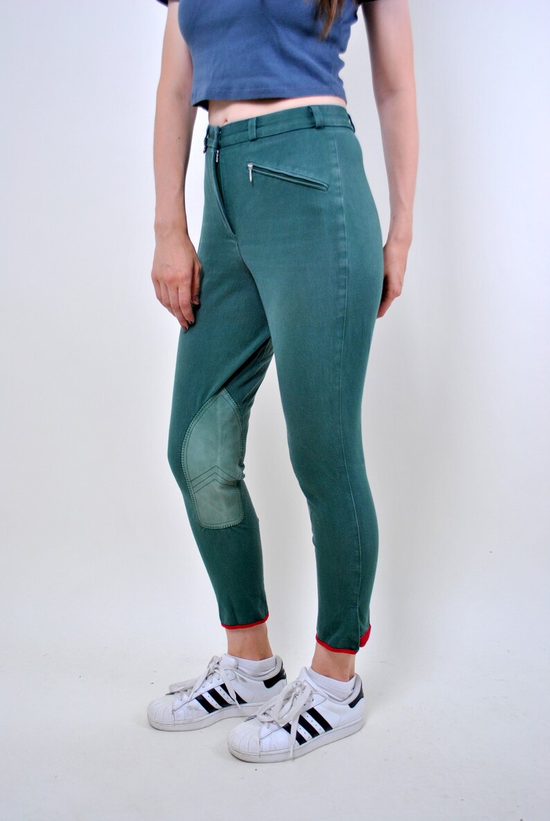 May include: Green riding pants with knee patches and red trim at the bottom. The pants are worn with white sneakers with black stripes.