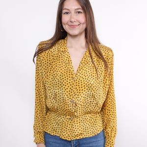 May include: A yellow blouse with a black dotted pattern. The long-sleeved top has a V-neck, button closure, and a cinched waist. The blouse is paired with blue jeans.