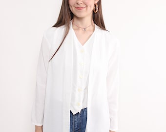 May include: A white long-sleeve shirt layered over a white button-up vest and blue jeans. The shirt has a draped front and a relaxed fit. The model is wearing gold hoop earrings and a necklace.