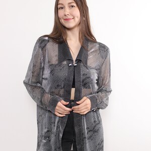 May include: A sheer, silver-gray patterned long-sleeve top with a collar and button closure. The top is worn over a black top and paired with black jeans. The fabric has a snakeskin-like design.