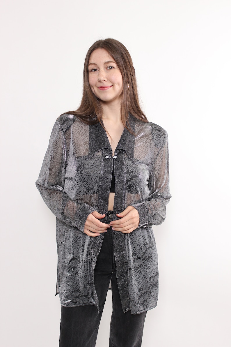 May include: A sheer, silver-gray patterned long-sleeve top with a collar and button closure. The top is worn over a black top and paired with black jeans. The fabric has a snakeskin-like design.