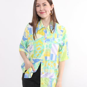 May include: A short-sleeved button-down shirt with a colorful geometric pattern in shades of yellow, blue, green, and purple. The shirt has a classic collar and yellow buttons. The model is wearing black jeans.