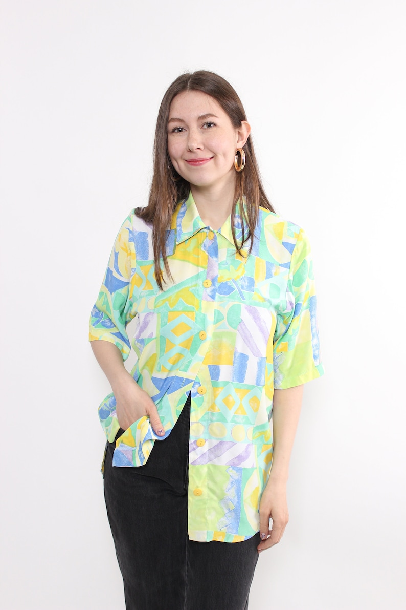 May include: A short-sleeved button-down shirt with a colorful geometric pattern in shades of yellow, blue, green, and purple. The shirt has a classic collar and yellow buttons. The model is wearing black jeans.
