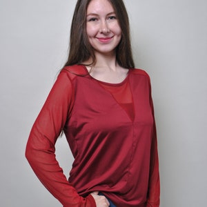 May include: A long-sleeved top in a deep red color featuring sheer mesh fabric on the sleeves and a V-shaped mesh panel at the neckline. The garment has a solid opaque red bodice and is paired with blue denim jeans, creating a layered appearance.