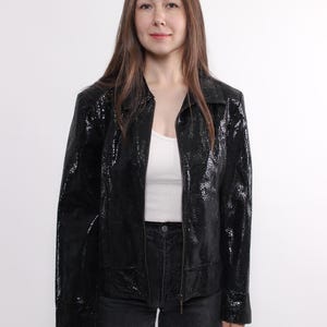 Peut inclure: Une veste en cuir noire avec une fermeture éclair et un motif peau de serpent. La veste a un col et des manches longues.