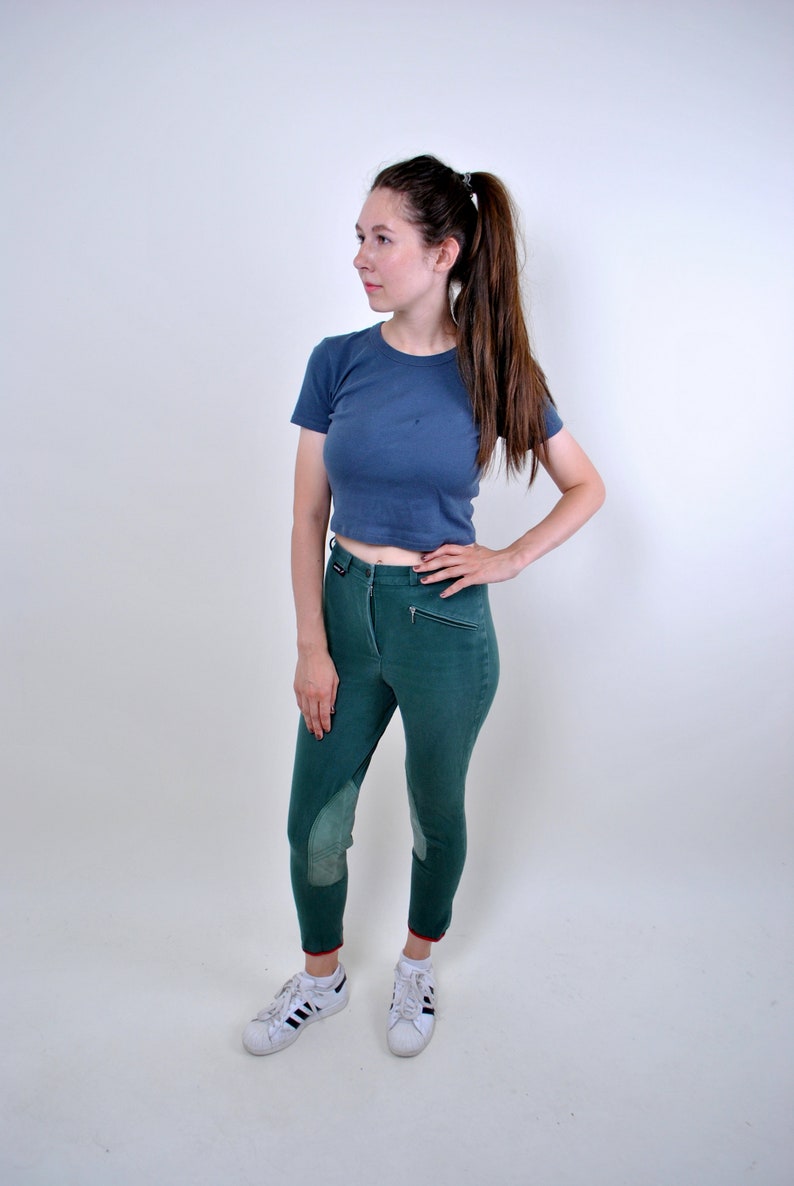 May include: A woman wearing a blue cropped t-shirt and green riding pants with knee patches. She is wearing white sneakers with black stripes.