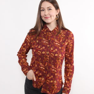 May include: A burgundy button-down shirt with a floral pattern in shades of gold, orange, and brown. The long-sleeved shirt has a classic collar and is paired with black pants. Gold hoop earrings complete the look.
