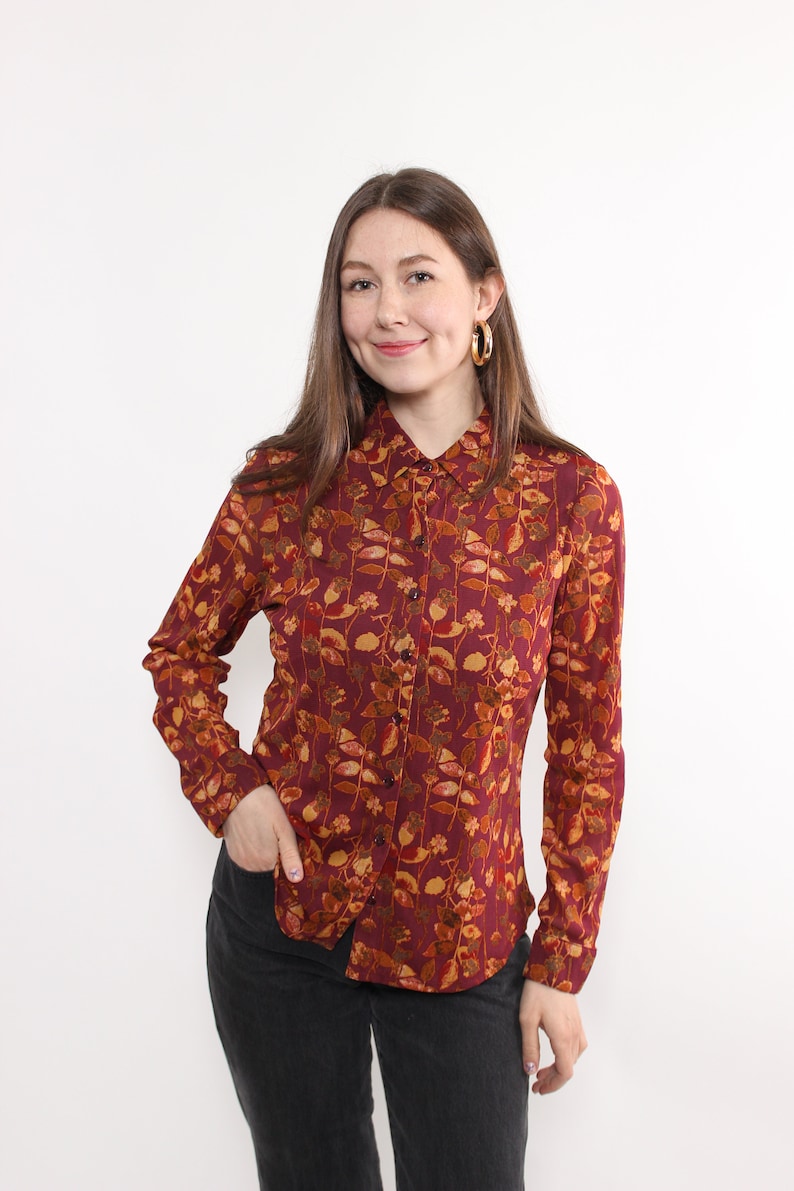 May include: A burgundy button-down shirt with a floral pattern in shades of gold, orange, and brown. The long-sleeved shirt has a classic collar and is paired with black pants. Gold hoop earrings complete the look.
