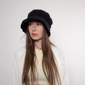 Könnte beinhalten: Ein schwarzer Cord-Eimerhut mit runder Krone und kurzem Rand. Der Hut wird von einer Person mit langen braunen Haaren, einem hellgelben Oberteil und einer weißen Strickjacke getragen. Der Hintergrund ist eine schlichte weiße Wand.