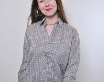 Può includere: Una donna indossa una camicia abbottonata a quadri grigi e gialli a maniche lunghe e una tasca sul petto. Indossa jeans blu e ha i capelli lunghi e castani.