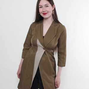 Puede incluir: Una chaqueta larga de manga larga, de color verde oliva y beige, con un panel beige en contraste y costuras blancas. La chaqueta tiene cuello, cierre de botón y bolsillos.