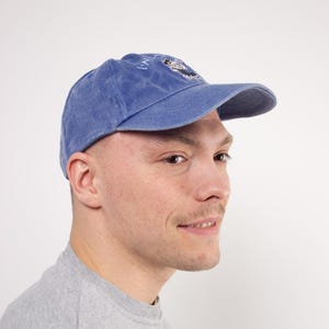 Könnte beinhalten: Eine blaue Baseballkappe mit gebogenem Schirm. Die Kappe hat ein gesticktes Design auf der Vorderseite. Die Kappe ist aus einem weichen, strukturierten Stoff gefertigt. Die Person trägt ein graues Sweatshirt.