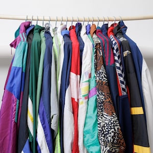 Könnte beinhalten: Eine Sammlung verschiedener Windbreaker-Jacken, die an einer Holzstange hängen. Die Jacken weisen verschiedene Farben auf, darunter Violett, Grün, Blau, Rot, Weiß und Schwarz, teilweise mit geometrischen Mustern, Streifen und abstrakten Drucken.