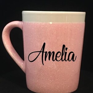 Może przedstawiać: Różowy kubek z brokatem, białym rantem i imieniem "Amelia" w czarnych literach.