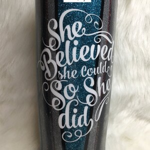 Op de afbeelding: Een turquoise, zwart en zilveren glitterbeker met een wit vinyl ontwerp met de tekst "She Believed She Could So She Did."