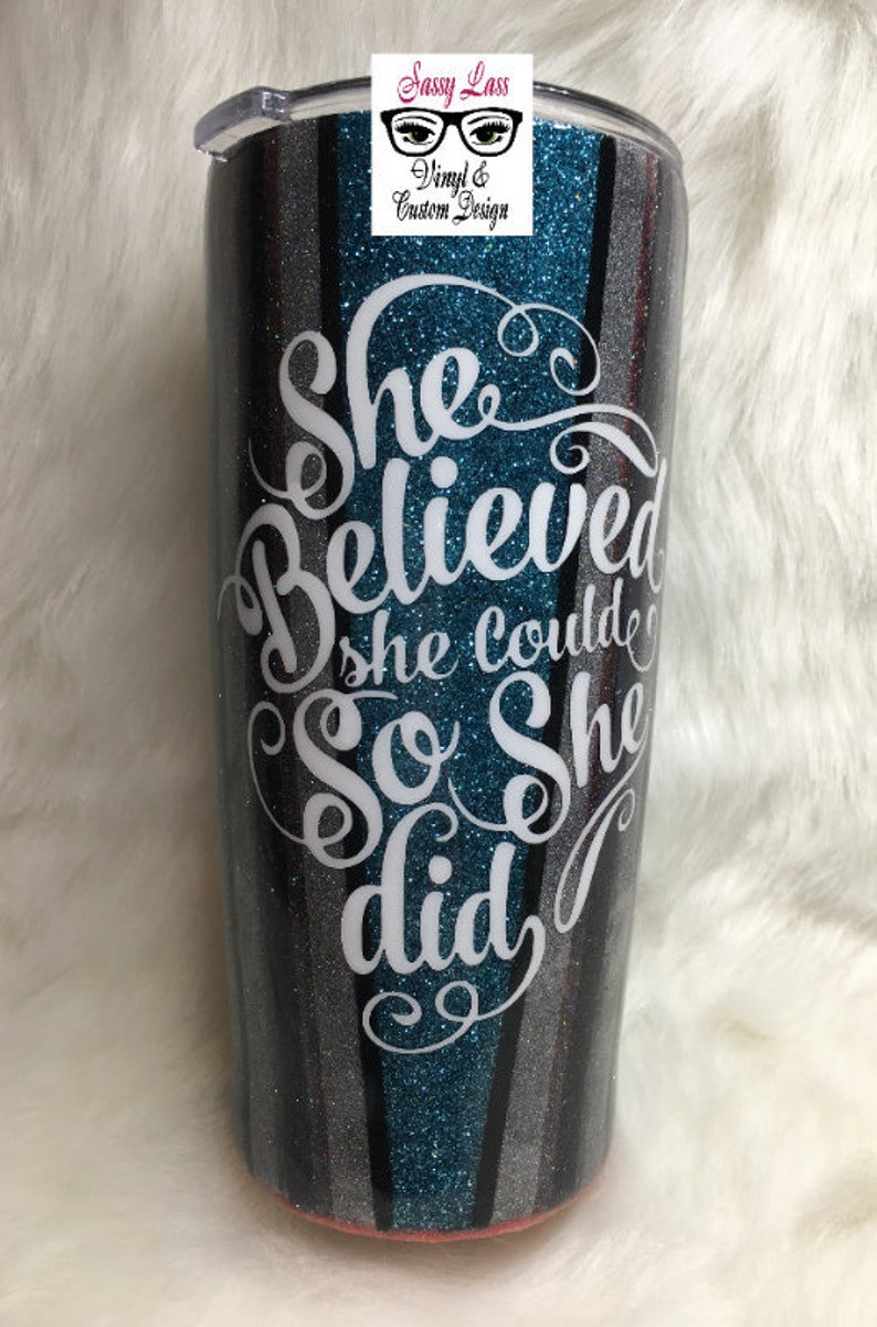 Op de afbeelding: Een turquoise, zwart en zilveren glitterbeker met een wit vinyl ontwerp met de tekst "She Believed She Could So She Did."