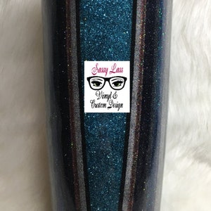 Op de afbeelding: Een turquoise, zwart, zilver en roze glitterbeker met verticale strepen. De beker heeft een doorzichtig deksel en een roze basis. De beker heeft een logo met de tekst "Sassy Lass Vinyl & Custom Design" met een bril en wimpers.