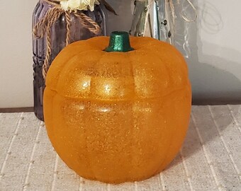 May include: An orange, glittery pumpkin-shaped container with a green stem sits in front of two vases of flowers. The vases are wrapped in twine and filled with colorful flowers, including pink, yellow, and white blooms. The background is a light gray wall.