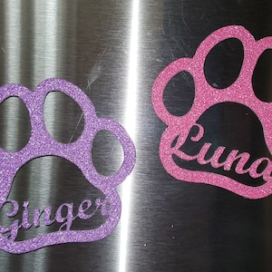 May include: Two glittery paw print magnets, one purple with the name "Ginger" and one pink with the name "Luna". The magnets are displayed on a metallic surface.