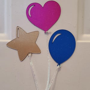 May include: A set of three decorative shapes including a glittery magenta heart, a glittery gold star, and a glittery blue oval balloon. Each shape features a white highlight detail and is attached to a thin, shimmering white ribbon string.