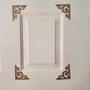 May include: Four decorative, glittery gold-colored corner brackets are affixed to a white cabinet door. Each bracket features an ornate, scrolled design. The brackets are evenly spaced around the corners of the door.