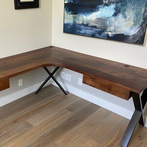 May include: An L-shaped desk made of dark brown wood, supported by black metal legs. The desk features a drawer and a circular cable management port. The desk is positioned in a corner.