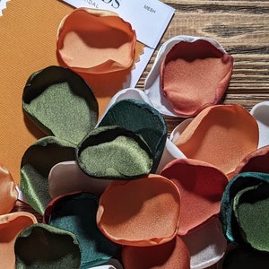 May include: Assortment of fabric flower petals in various colors, including green, orange, and white. The petals are arranged on a wooden surface.