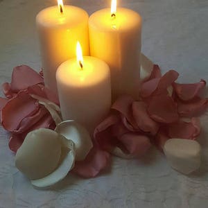 May include: Three white pillar candles are lit and surrounded by pink rose petals on a white tablecloth.