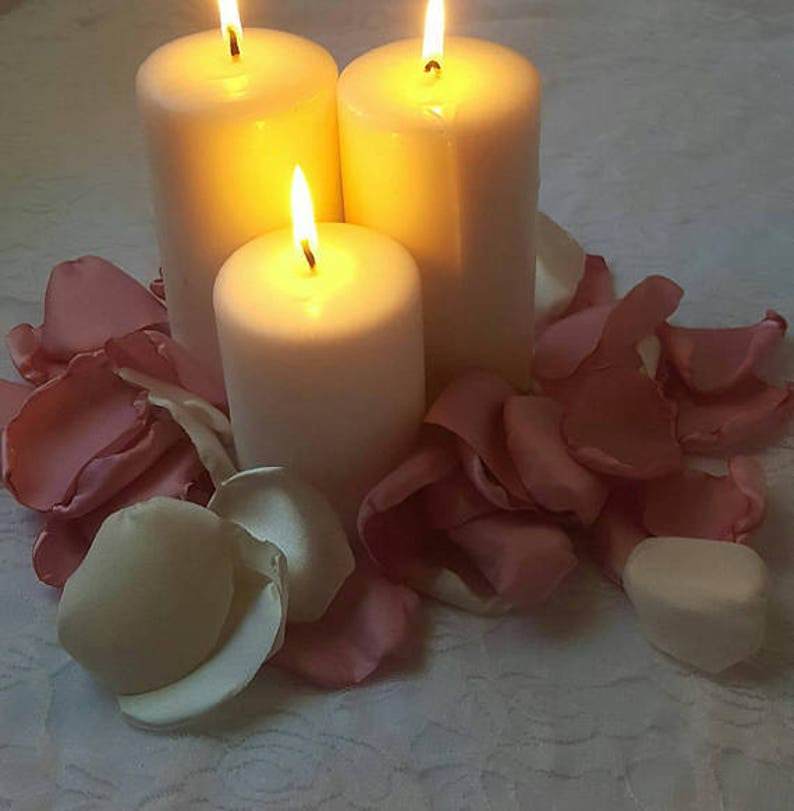 May include: Three white pillar candles are lit and surrounded by pink rose petals on a white tablecloth.
