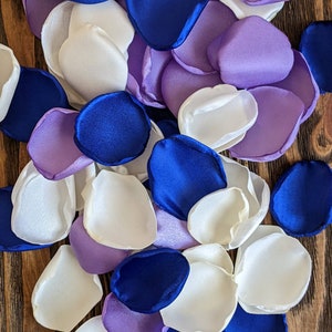 Può includere: Assortimento di petali di fiori di seta nei colori bianco, lavanda e blu reale. I petali sono sparsi su una superficie di legno.