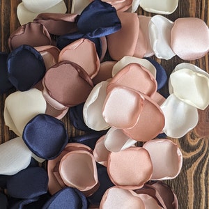 May include: A pile of silk flower petals in shades of ivory, blush pink, and navy blue. The petals are arranged in a random pattern and are perfect for wedding decorations or other special events.