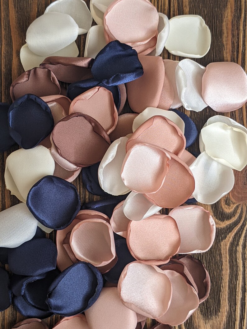 May include: A pile of silk flower petals in shades of ivory, blush pink, and navy blue. The petals are arranged in a random pattern and are perfect for wedding decorations or other special events.
