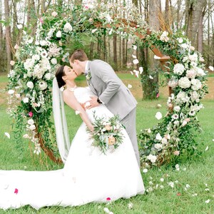 May include: A bride and groom kiss in front of a floral archway decorated with white and pink flowers. The bride is wearing a white wedding dress and the groom is wearing a gray suit. White flower petals are falling from the sky.