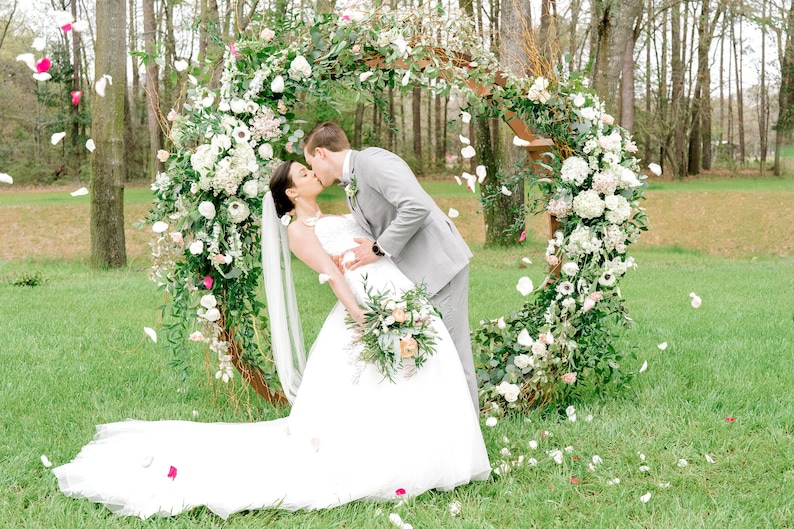 May include: A bride and groom kiss in front of a floral archway decorated with white and pink flowers. The bride is wearing a white wedding dress and the groom is wearing a gray suit. White flower petals are falling from the sky.