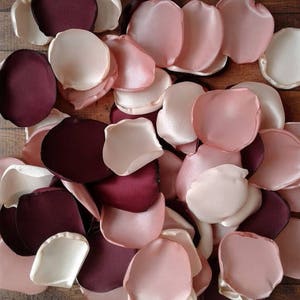 May include: A collection of burgundy, blush pink, and ivory satin rose petals. The petals are arranged in a random pattern and are perfect for wedding decorations or other special events.