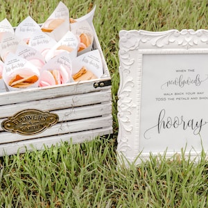May include: A white wooden box filled with small white cones containing pink flower petals. A white sign with black calligraphy reads "When the newlyweds walk back your way toss the petals and shout hooray".