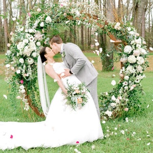 May include: A bride and groom kiss in front of a floral archway decorated with white and pink flowers. The bride is wearing a white wedding dress and the groom is wearing a gray suit. White flower petals are falling from the sky.