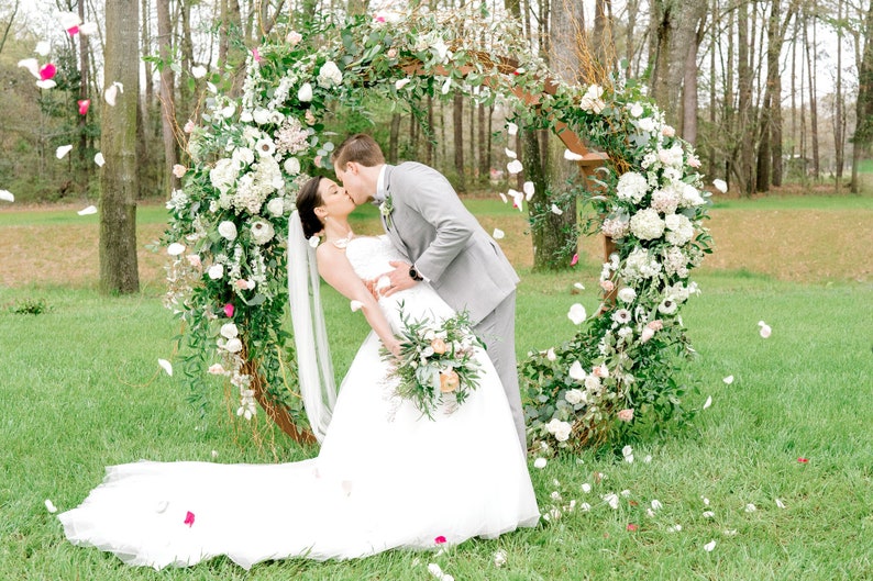 May include: A bride and groom kiss in front of a floral archway decorated with white and pink flowers. The bride is wearing a white wedding dress and the groom is wearing a gray suit. White flower petals are falling from the sky.