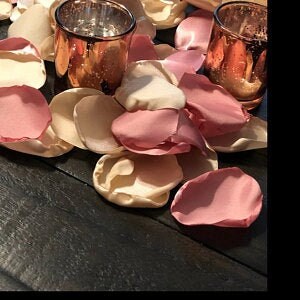 May include: A pile of rose petals in pink, cream, and ivory colors on a dark wood surface. Two copper-colored glass candle holders are in the background.