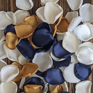 Könnte beinhalten: Ein Haufen weißer, goldener und marineblauer Seidenblumenblätter. Die Blütenblätter sind auf einer Holzoberfläche verstreut.
