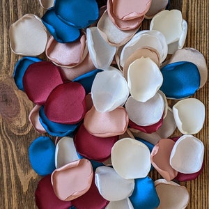 Op de afbeelding: Een verzameling zijden bloemblaadjes in tinten bordeauxrood, blos roze, wit en teal blauw. De bloemblaadjes zijn op een houten oppervlak in een stapel gerangschikt.